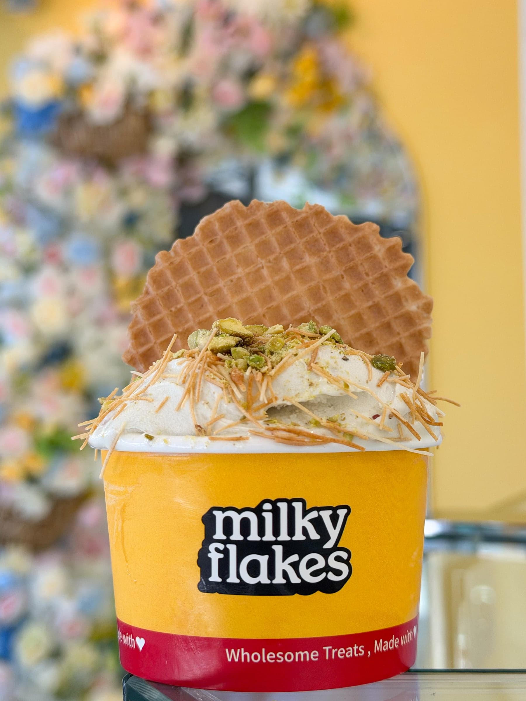 A cup of soft scoops topped with pistachio, a waffle standing behind it, at the shop's flower wall.